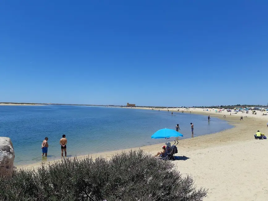 Plage de sable calme avec eau bleue et promeneurs, parasol bleu et vacanciers près du bord; maisons de pêcheurs au loin.