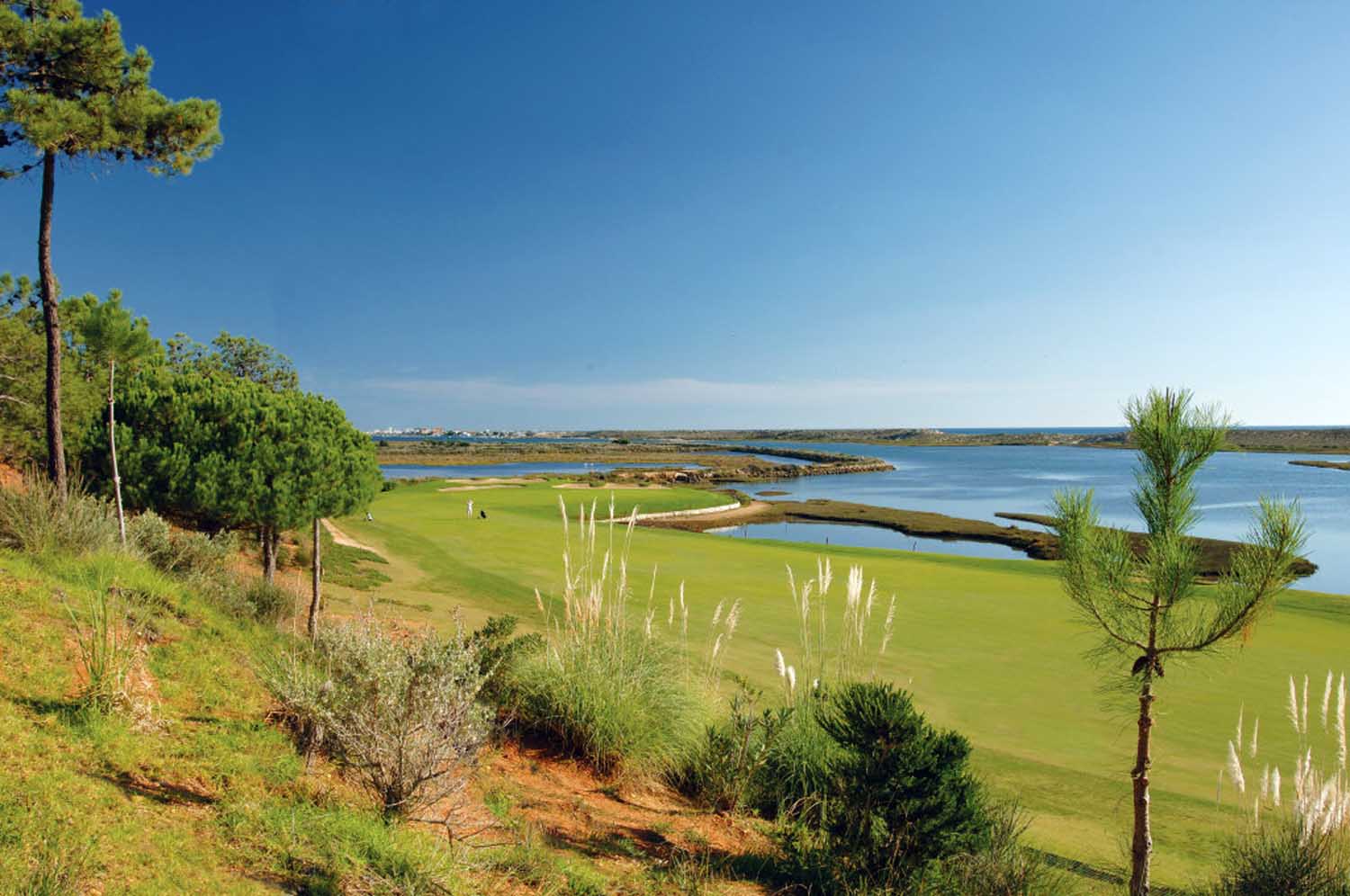 Lush coastal golf course with a green fairway along the sea, surrounding shrubs and trees under a clear blue sky. Perfect for t to play golf in algarve.