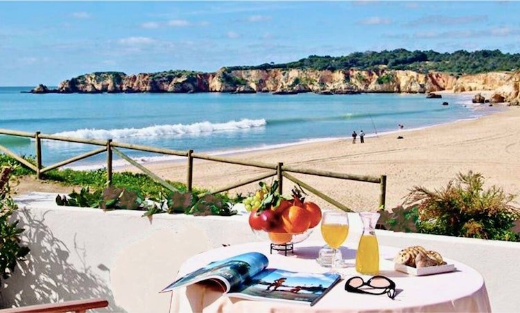 Table à café face à une plage dorée, avec magazine ouvert, fruits et boissons, vue mer et rochers en arrière-plan. villa de luxe portugal algarve idéale.