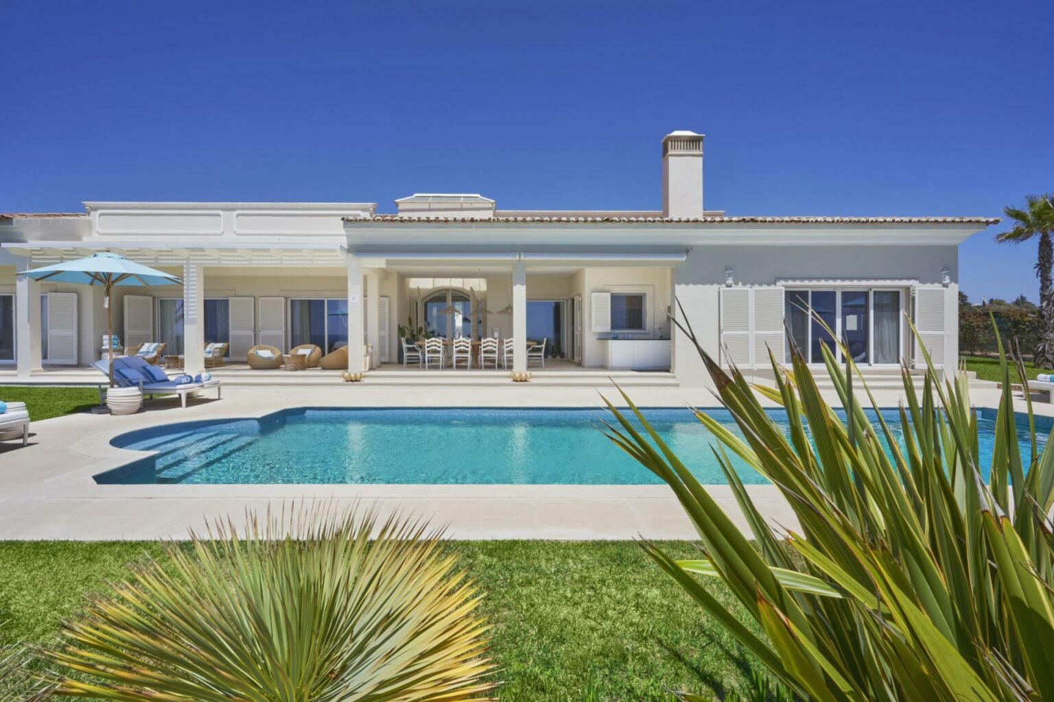 Villa de luxe Portugal Algarve avec piscine privée et terrasse bahutage, façade blanche lumineuse et espaces de détente, verdure et palmier en avant.