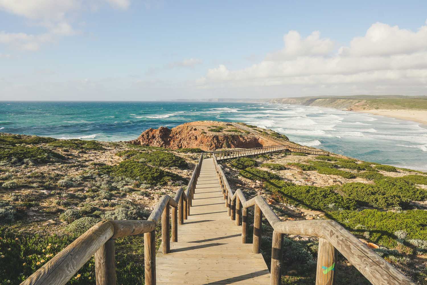Wooden boardwalk stairs descending toward a rugged coastline with blue Atlantic waves, green scrub, and a distant horizon under a partly cloudy sky. golf algarve weather conditio