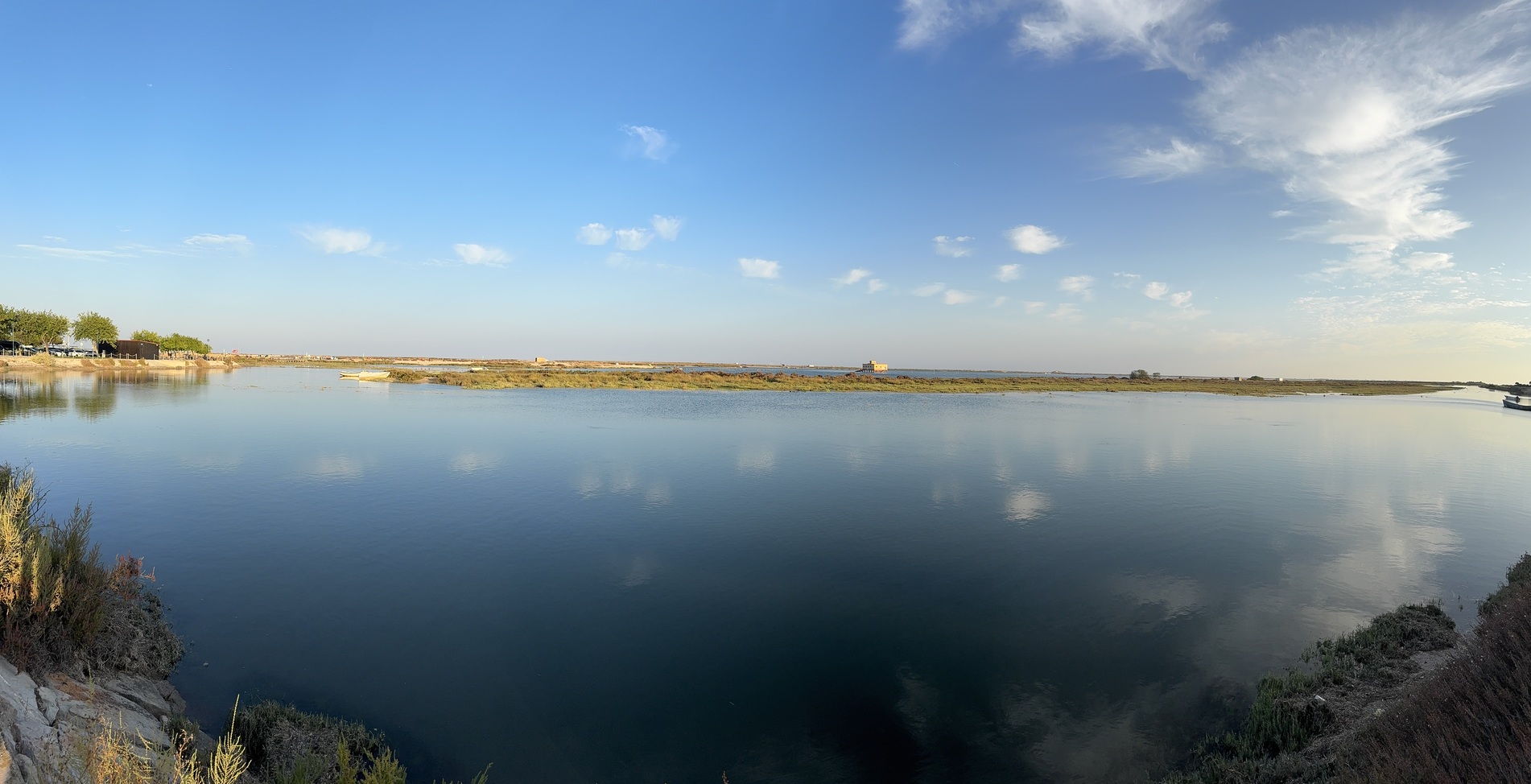 Tranquil coastal inlet at sunset with calm blue water, a distant shoreline, and a clear sky reflected on the surface. Sea views and serene ambience.