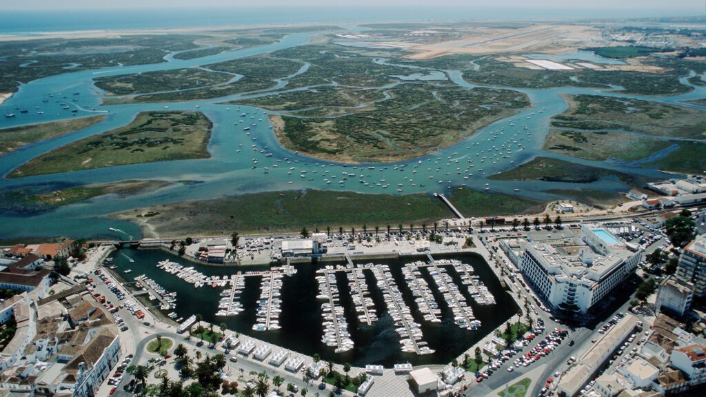 Aerial view of a marina with boats and lush wetlands forming a winding estuary; coastal town in Algarve, Portugal (Ria Formosa).