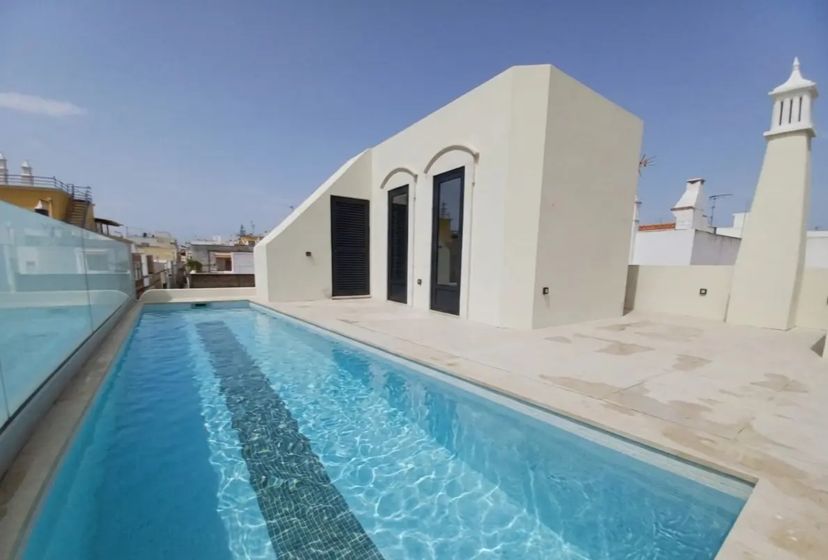 Piscine longue sur terrasse d’une maison blanche de charme, façade moderne avec grandes portes vitrées et vue partielle du village de Fuseta.
