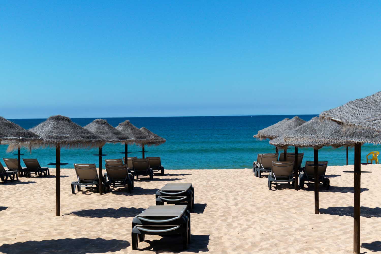 Plage en Algar­ve avec des parasols en paille et transats, mer turquoise et ciel bleu clair. algarve climat mai intégré.