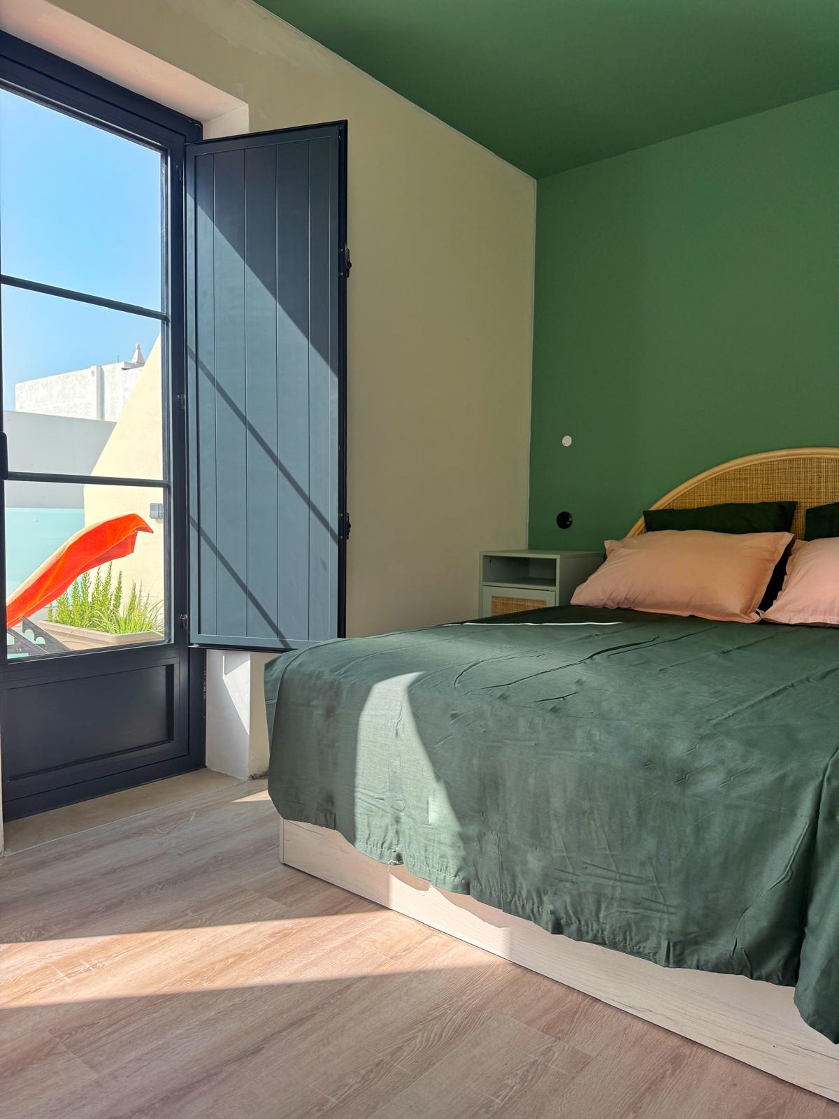 Bright bedroom with a dark green bedspread, pink cushions, and a wicker headboard. Large glass door with navy shutters leads to outside, letting sunlight onto light wooden flooring.