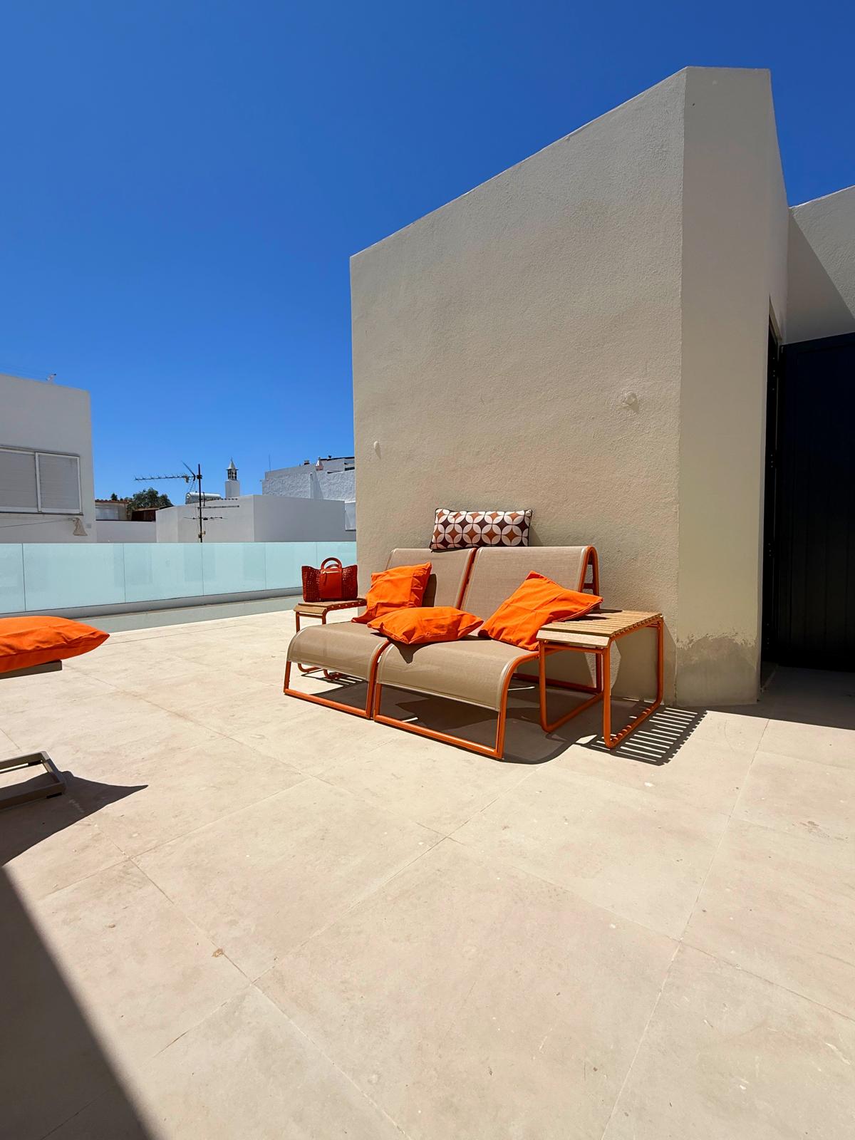 Luxurious sunlit terrace with beige outdoor seating and orange cushions, modern white walls, and clear blue skies. What personalized guest services do high-end Algarve rentals offer?