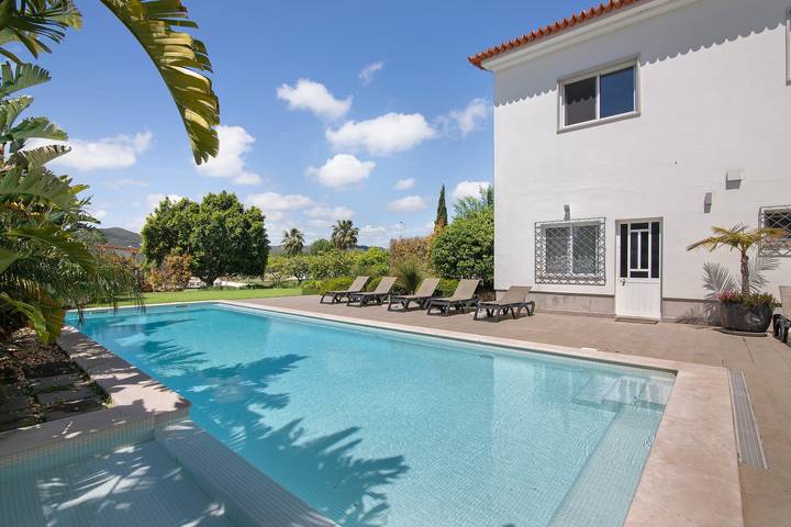 Poolside view of a white two-storey villa with loungers, lush garden, and clear blue sky in a sunny Algarve setting. Best alternatives to airbnb for upscale Algarve villa rentals.