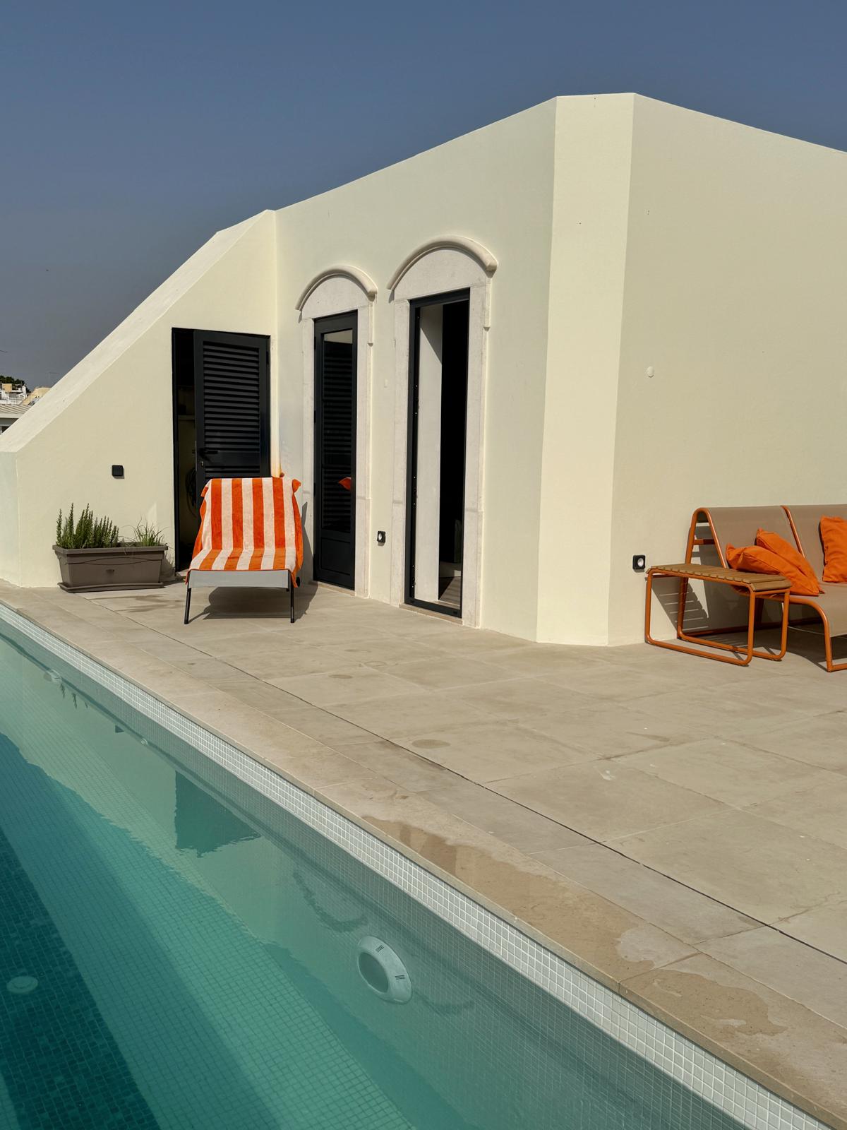 Rooftop terrace with a white corner building, two arched doorways, striped orange chair, orange-cushioned lounge, and a turquoise pool along the edge. What personalized guest services do high-end Algarve rentals offer?
