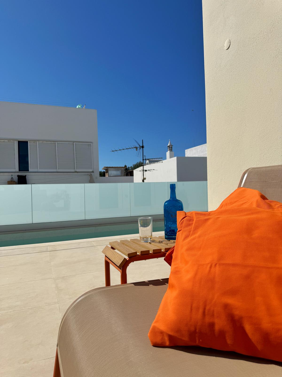 Sunlit Algarve terrace with a beige sunbed and bright orange cushion, a wooden side table, blue bottle and glass by a private pool, modern white buildings in the background. what to look for in a high-end holiday rental in southern portugal?