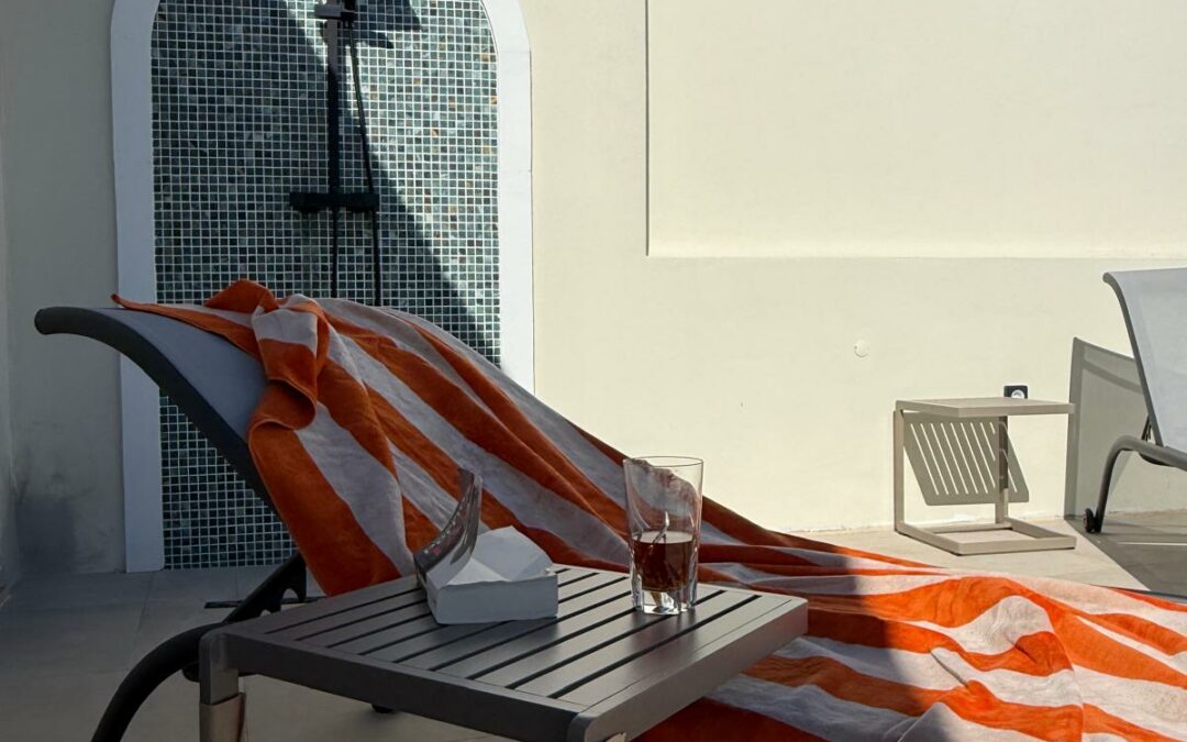 Sunlit rooftop terrace with a reclining lounge chair draped in an orange and white striped towel, a small table holding a glass and tissues, against a white wall and a tiled outdoor shower.
