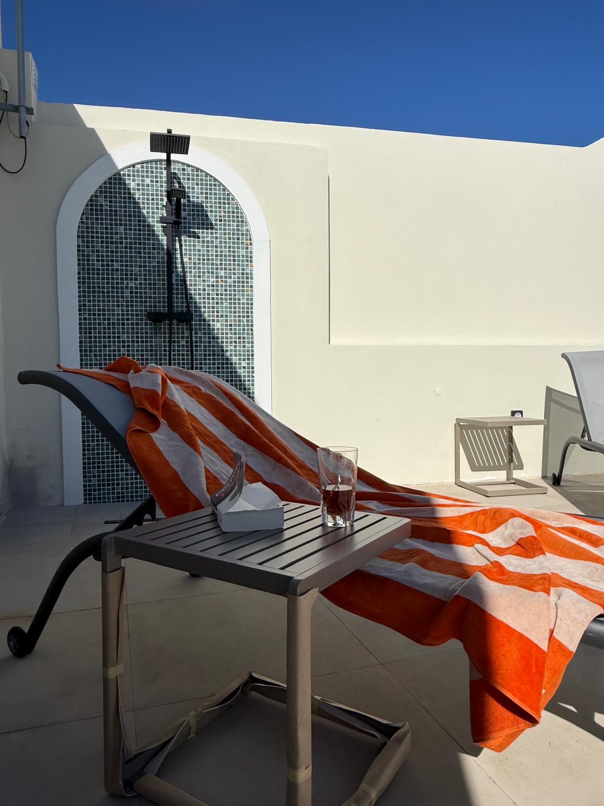 Sunlit rooftop terrace with a reclining lounge chair draped in an orange and white striped towel, a small table holding a glass and tissues, against a white wall and a tiled outdoor shower.