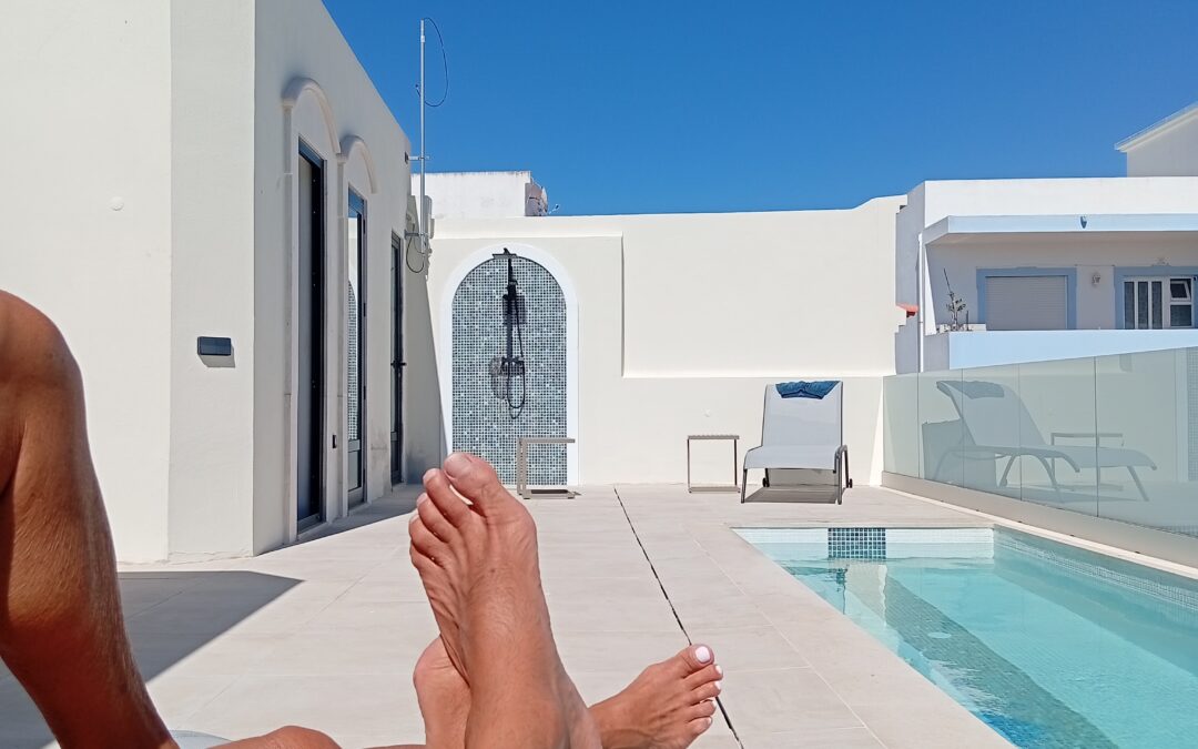 Sun loungers by a bright white villa with a rectangular pool under a clear blue sky. Sunbathers’ legs in the foreground.